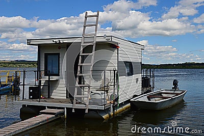 White square shaped house boat with ladder leaning on front wall and wooden bridge in front Editorial Stock Photo