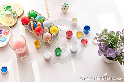 On a white table, in daylight, there are brushes in a glass of water, multi-colored eggs in a tray, jars of paint. Stock Photo