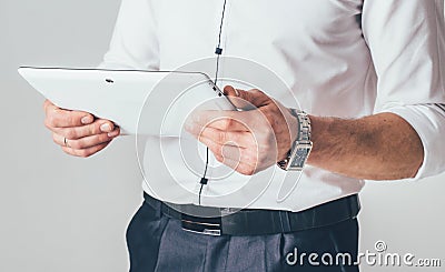 A white tablet is in the hands of a man. He stands in a white shirt and black trousers and reads information from the gadget Stock Photo