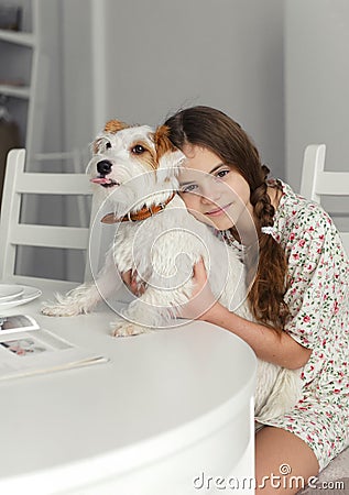 Teenage girl 10 years old sits at the kitchen table and hugs jack Russell`s white furred dog Stock Photo