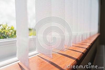 White vertical window blinds slats with cordless glued weighted pockets on the end casting shadows on the wooden window sill Stock Photo
