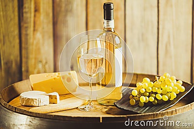 White wine in glass, bottle with grape and cheese on barrel front wood wall background. Stock Photo