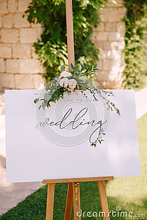 White wooden easel with an inscription on the background of a stone wall and greenery. Caption: Welcome to our wedding Stock Photo