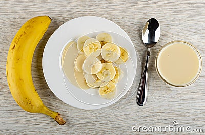 Whole banana, white plate with slices of banana and condensed milk, spoon, bowl with milk on wooden table. Top view Stock Photo