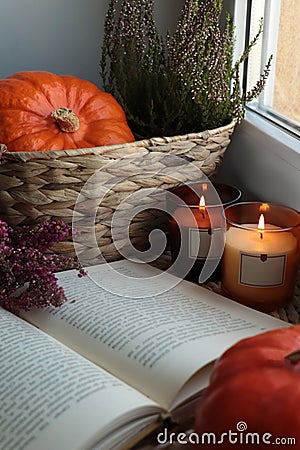 Wicker basket with beautiful heather flowers, pumpkin, burning candles and open book near window indoors Stock Photo