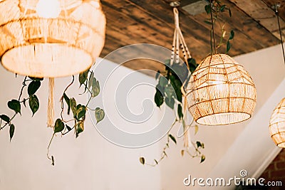 Wicker handmade chandelier lamps on the wooden ceiling, hanging flower pots with green plants on the white wall Stock Photo