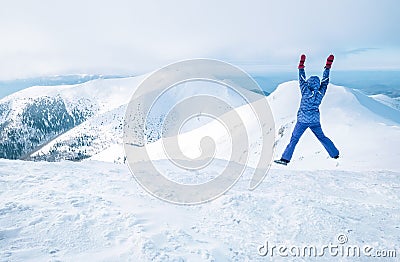 Wide angle shot of woman dressed in ski warm clothing jumping on the mountain peak with snowy range and valley on the background. Stock Photo