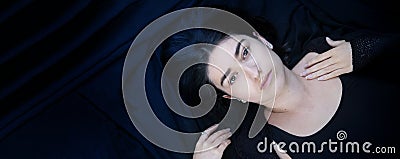 Wide banner panorama portrait of black haired young lying woman with beguiling hypnotic look on blue black fabric background, Stock Photo