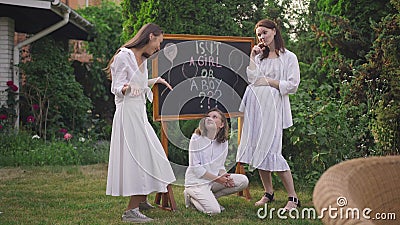 Wide shot cheerful thoughtful pregnant woman and girls standing at blackboard with Is it boy or a girl making confused stock footage Wide shot cheerful thoughtful pregnant woman and girls standing at blackboard with Is it boy or a girl making confused stock footage
