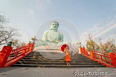 Wide shot and side view of young Asian monk hold red umbrella and walk down on stair in front of green big buddha statue with soft Stock Photo
