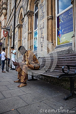 A black man wearing traditional african dress sitting on a bench looking at his mobile phone Editorial Stock Photo