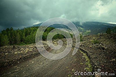 Windirg dirt road along the slopes of Etna Mount leading to the tree line under stormy sky Stock Photo