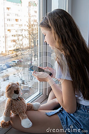 On the windowsill sits a cute teenage girl with a soft toy and a phone. The sun is shining outside the window. Stock Photo