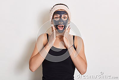 Winsome girl treating and caring for her skin. Woman with hairband on head and facial mask looking at camera over white background Stock Photo