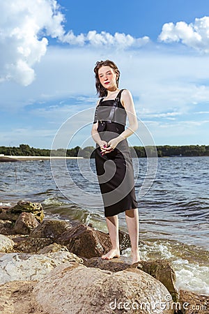 Winsome Relaxing Caucasian Brunette Girl Posing in Black Dress On Stone Line At Sea During Sunny Day Outdoors Stock Photo