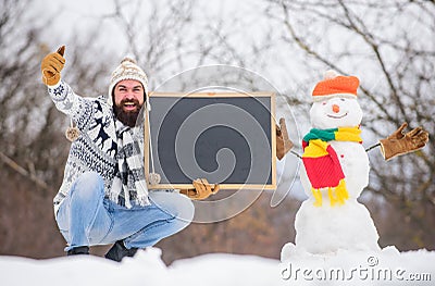 Winter announcement. Hipster knitted hat and gloves show blackboard. Winter holidays. Guy and snowman snowy nature Stock Photo
