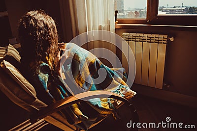 Winter and Christmas holidays concept. Young woman sitting in comfortable modern chair near radiator with mug of tea wrapped in wa Stock Photo