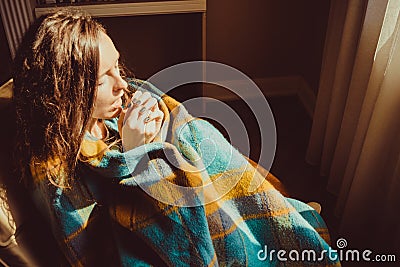 Winter cold concept. Young freezing woman in comfortable chair breathe warm air on frozen hands wrapped in warm fluffy woollen pla Stock Photo