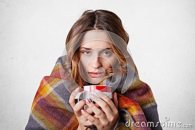 Winter cold sickness concept. Freezing beautiful woman wrapped in warm checkered plaid blanket, drinks hot beverage, tries to warm Stock Photo