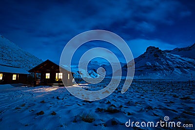 Winter image of a wooden house with light from the window. Landscape with moonlight on the snow with mountains in the background. Stock Photo