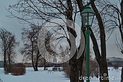 Winter park with green lantern, bare trees and bushes. Background of embankment of Saint Petersburg with buildings and St. Isaac C Stock Photo