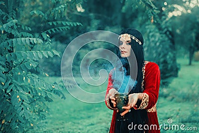 Wise fortune-teller in silk red scarlet dress and gold decoration stands alone in forest with magic bowl, calling Stock Photo