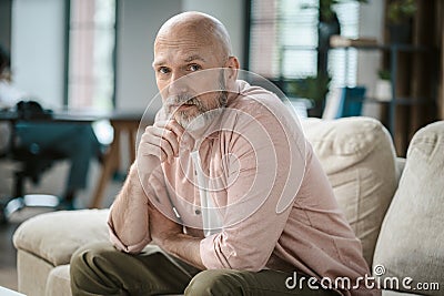 Wise senior man, with touch of grey in beard, looks directly into camera within cozy interior of home. His hand gently Stock Photo