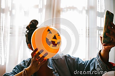 Witch holding a pumpkin balloon with her hand, covering her face and taking a photo with mobile phone. Young woman celebrating Stock Photo