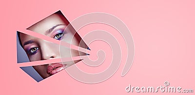 Woman with beautiful bright makeup and pink lipstick looks through triangular slits in pink paper. Advertising cosmetics Stock Photo