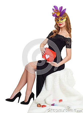 Woman in black evening gown and carnival mask with gift box. Sit on white fur. Valentine holiday and party concept. Stock Photo