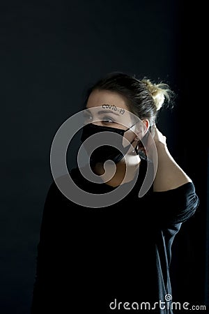 Woman in a black protective mask with the words covid-19 written on her forehead. Girl pushes a lock of hair from her Stock Photo