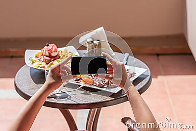 Woman blogger taking a picture on mobile phone plate with lamb potato and salad on the table outside on the balcony. Top view. Stock Photo