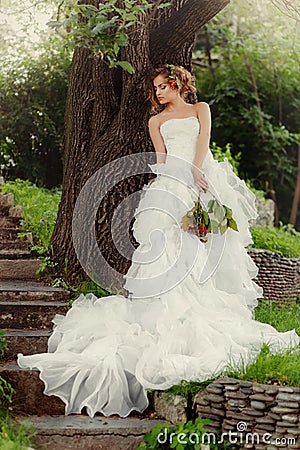 Woman bride in a wedding dress relaxes standing by a large tree. Stock Photo