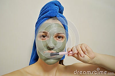 A woman with a brush applies a clay mask to her face with blue towel on her head Stock Photo