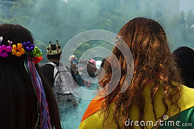 Woman carrying rainbow flag on her back next to woman with all rainbow flowers amid blue green haze and crowds of people at the LG Editorial Stock Photo