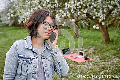 A woman with children looks at a grazing cow on the background of a blooming garden and green grass. Blanket with laptop and Stock Photo