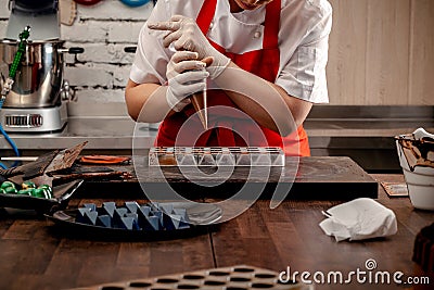 A woman confectioner with red uniform and white sterile gloves do a set of colorful chocolates from milk chocolate on a Stock Photo