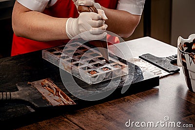 A woman confectioner with red uniform and white sterile gloves do a set of colorful chocolates from milk chocolate on a Stock Photo