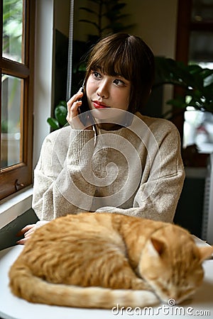 A woman in cozy sweater talking on the phone while her cat is sleeping on a table Stock Photo