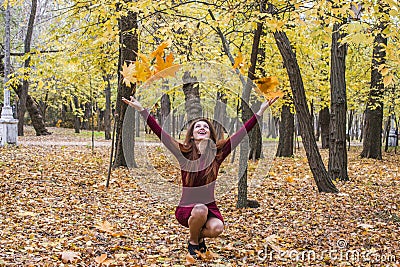 The woman crouched in the park in the fall and throws up yellow leaves. Stock Photo