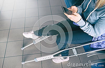 Woman on crutches, leg is splinted, siting in waiting chair in hospital and using smartphone. Knee brace support, bandaged leg Stock Photo