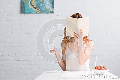 woman with cup of coffee and bowl of strawberries covering face with book Stock Photo