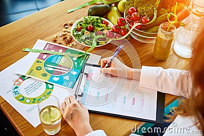 Woman dietitian in medical uniform with tape measure working on a diet plan sitting with different healthy food ingredients in the Stock Photo