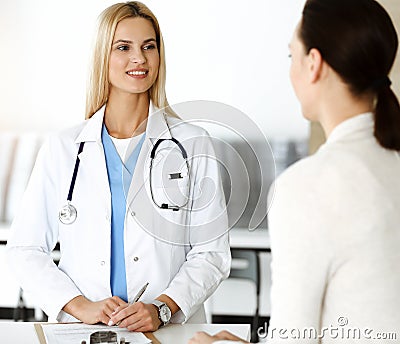 Woman-doctor at work in sunny hospital is happy to consult female patient. Blonde physician checks medical history Stock Photo