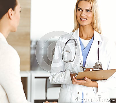 Woman-doctor at work in sunny hospital is happy to consult female patient. Blonde physician checks medical history Stock Photo