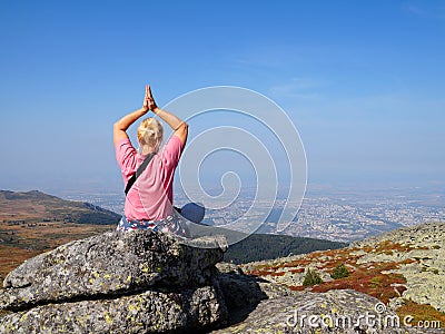 A woman does yoga on the top of a mountain, at the foot of which there is a large metropolis Stock Photo