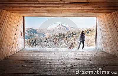 Woman with dog stay in big wooden hangar and looks on snowy mountains Stock Photo