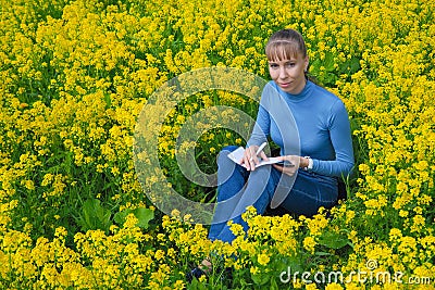 Woman draws sitting on a grass in the daytime. She paints sitting on a rapeseed field. Stock Photo
