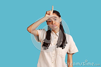 Woman with dreadlocks makes loser gesture over forehead, being disrespectful, looking at camera. Stock Photo