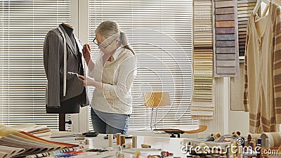 Woman dressmaker working with tailoring mannequin, sews the buttons to the jacket. With colorful clothes, fabrics and tools on the Stock Photo